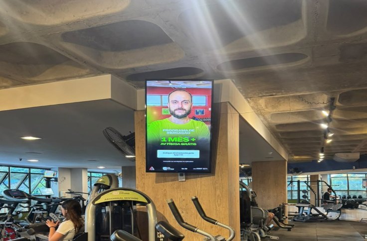 Tela vertical instalada no piso da musculação da Academia Via Campus exibindo conteúdo publicitário